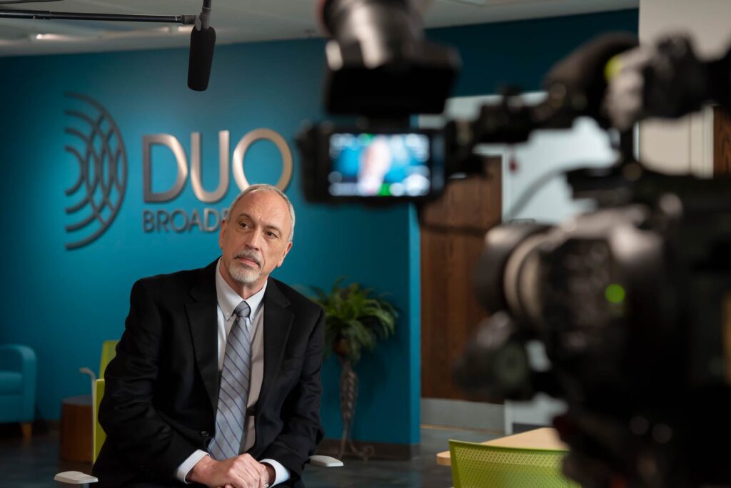 TV Camera pointing at man in front of a wall with the DUO Broadband logo.