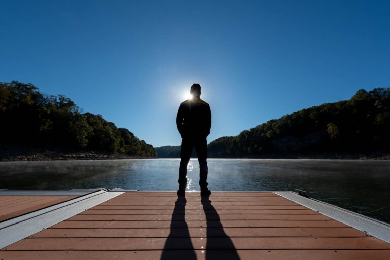 Man standing on edge of a dock on the lake silhouetted against the sun.