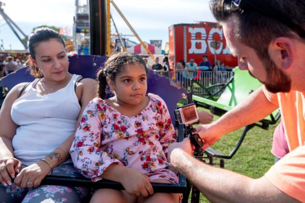 Videographer attaching GoPro camera to carnival ride chair with mother and daughter looking on.