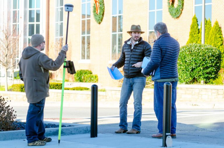 Three people on sidewalk with survey equipment and signage location maps.