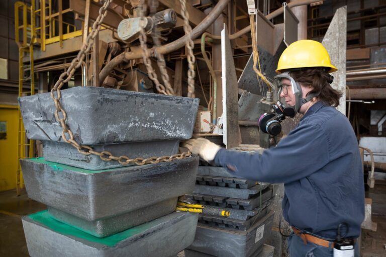 Worker in hard hat and respirator face shield attaching chain to large block of lead in factory environment.