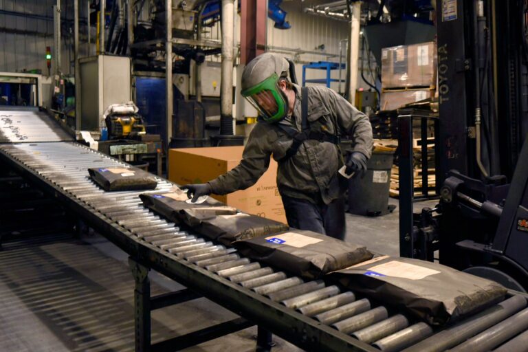 Factory worker handling product packages on conveyor belt in personal protective gear.