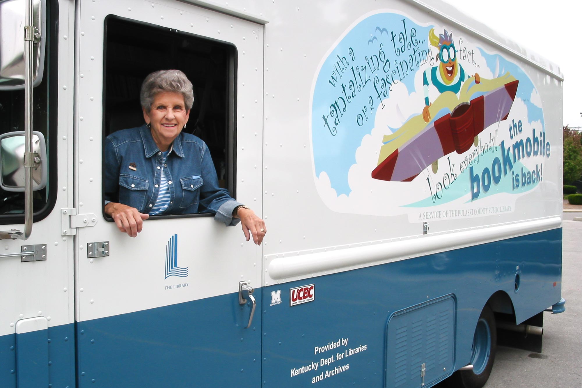 Bookmobile with side graphics of acartoon character riding an open book in the sky.
