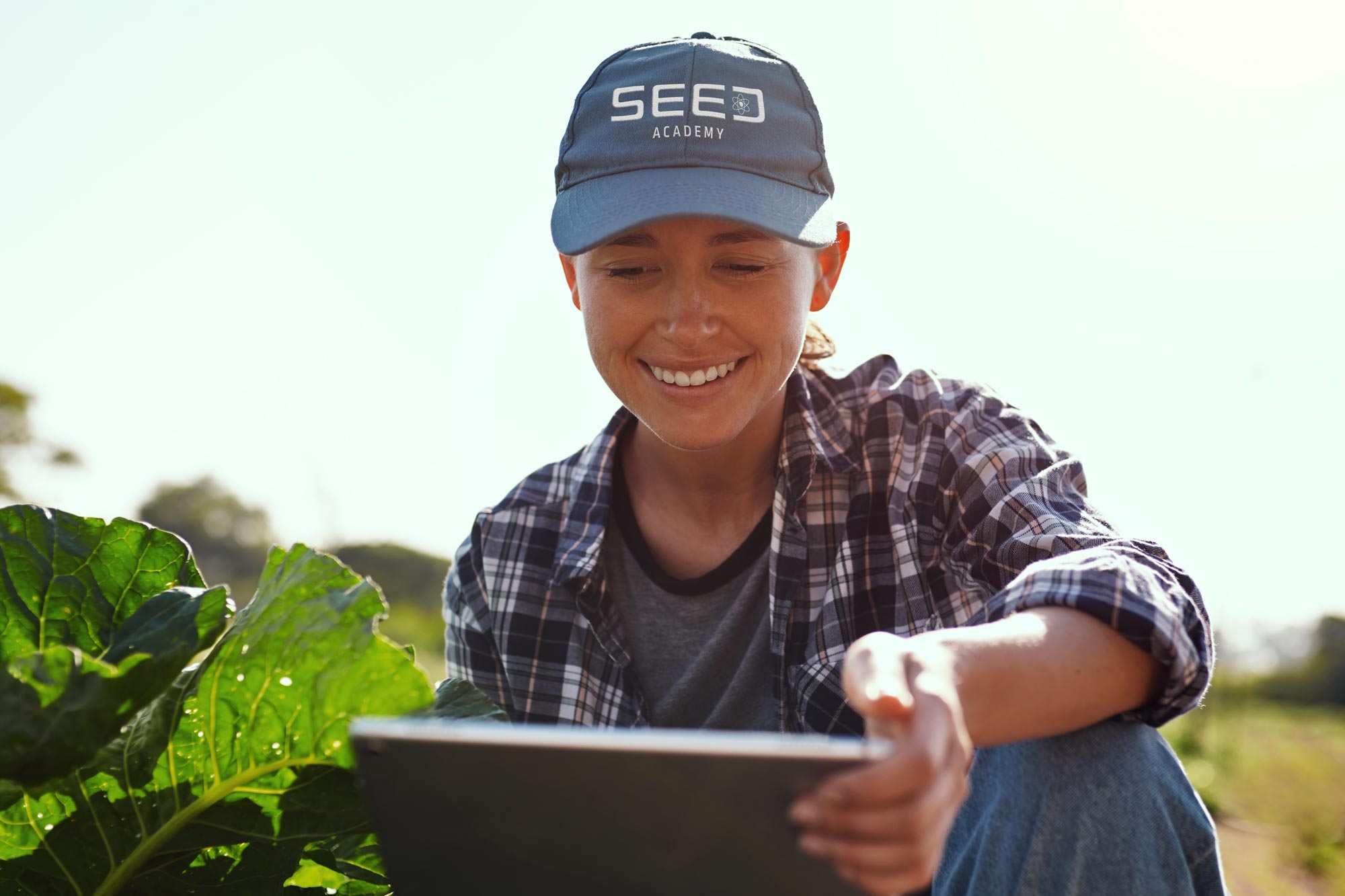 Young woman holding an iPad in a farm field waring a cap with "SEED ACADEMY" logo