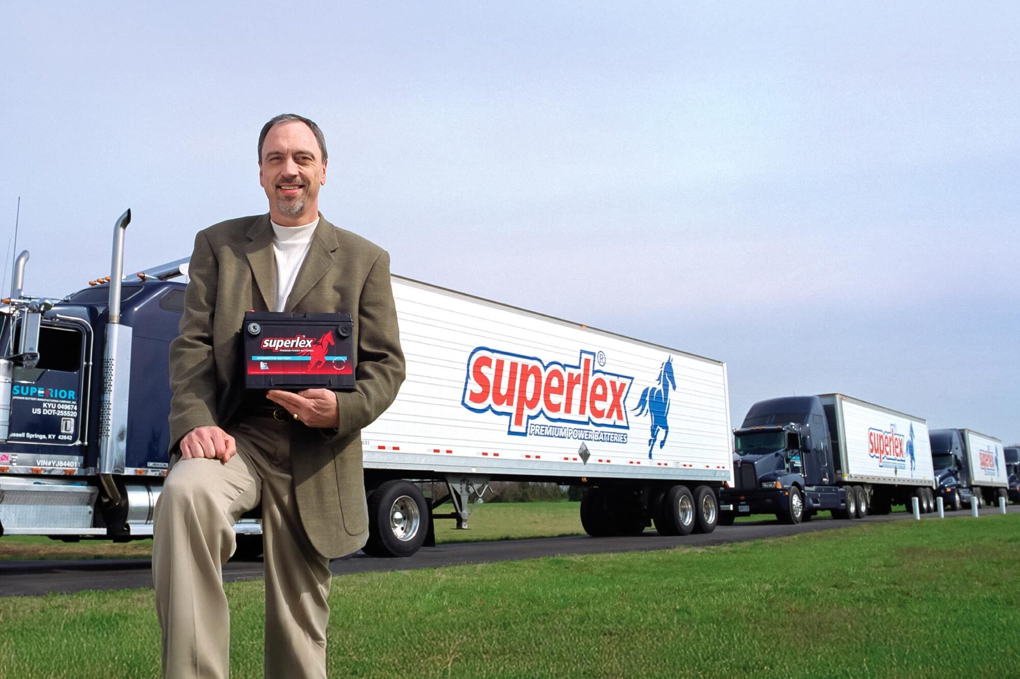 Man holding a car battery with row a trucks in the background with large Superlex Premium Power Batteries logo on the side of the trailers