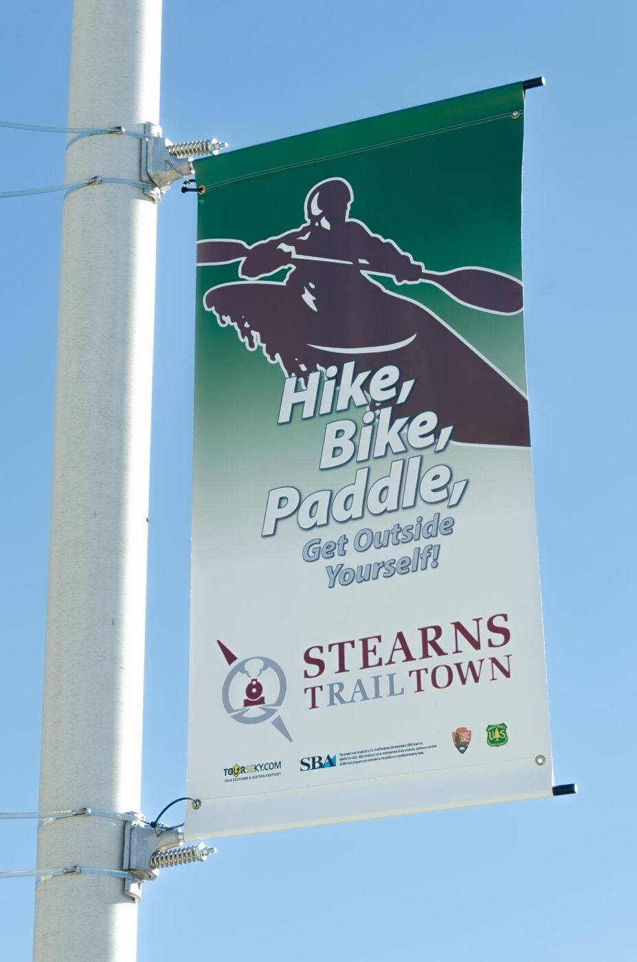 Banner on pole with a stylized kayaker that says Hike, Bike, Paddle, Get Outside yourself with Stearns Trail Town logo