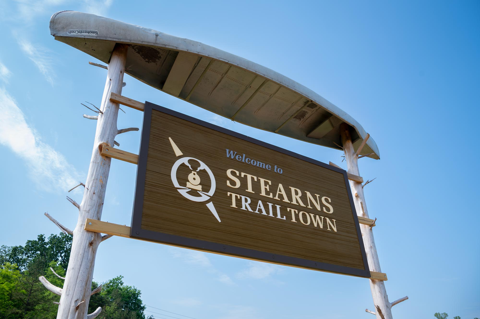 Laarge sign "Welcome to Stearns Trail Town" with logo on rustic poles with a canoe upside down on top