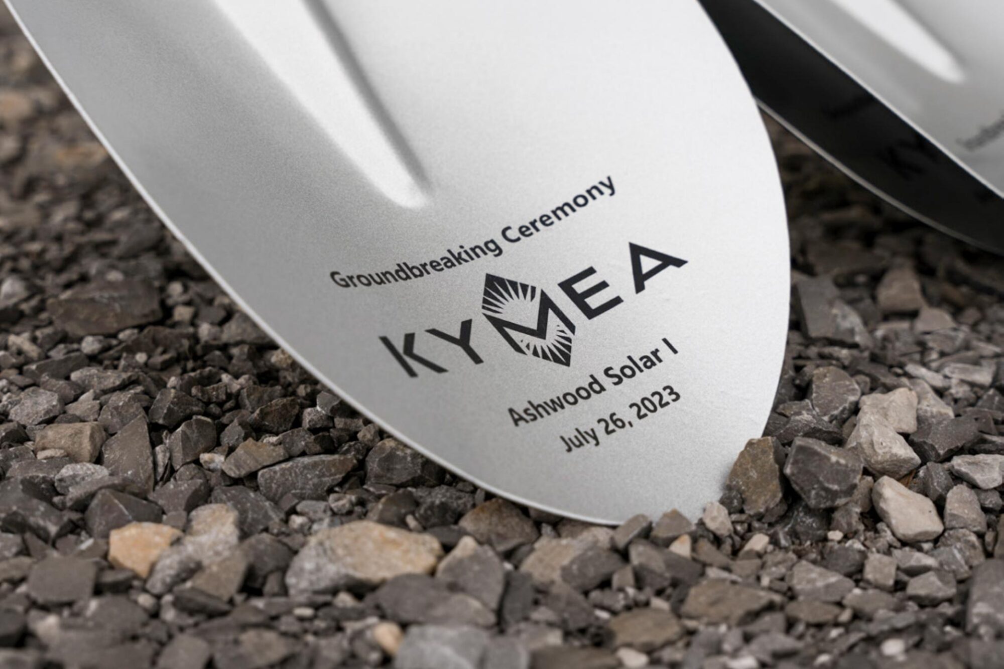 Shovel head touching a gravel bed at a ground breaking with the KYMEA logo and "Groundbreaking Ceremony" - "Ashwood Solar 1, July 26, 2023"
