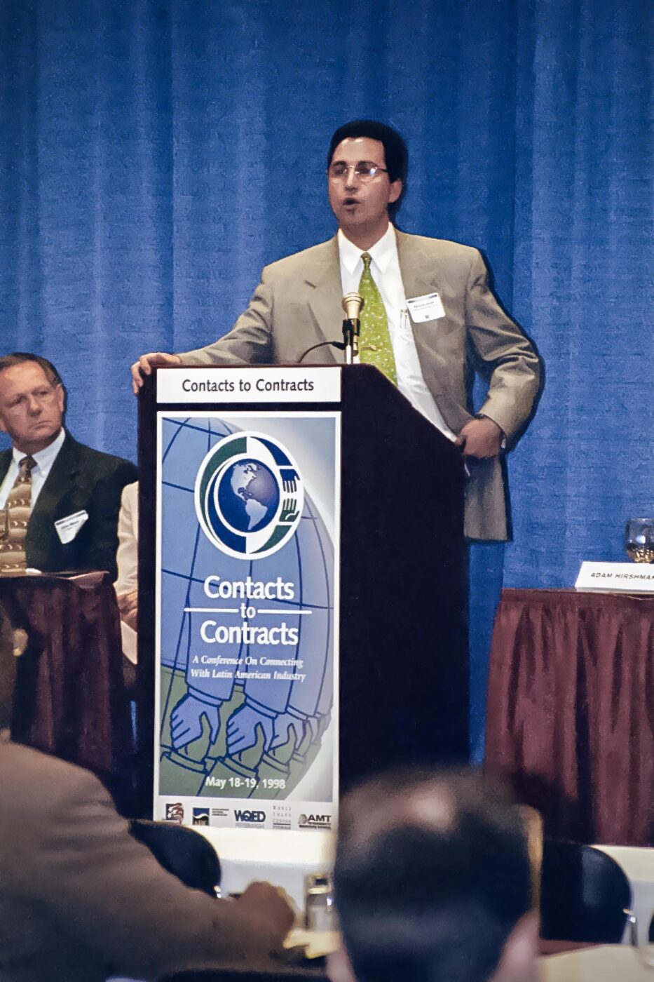 Speaker at a podium with the Contracts to Contacts logo on the front.