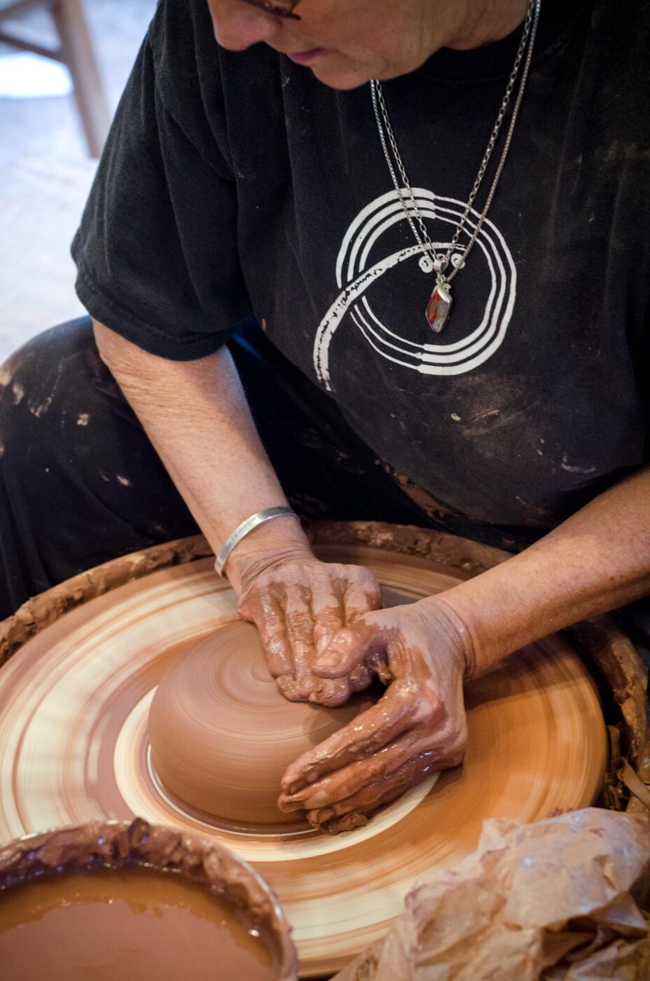 Potter trowing clay pot on a wheel with Tater Kob Pottery symbol on their t-shirt