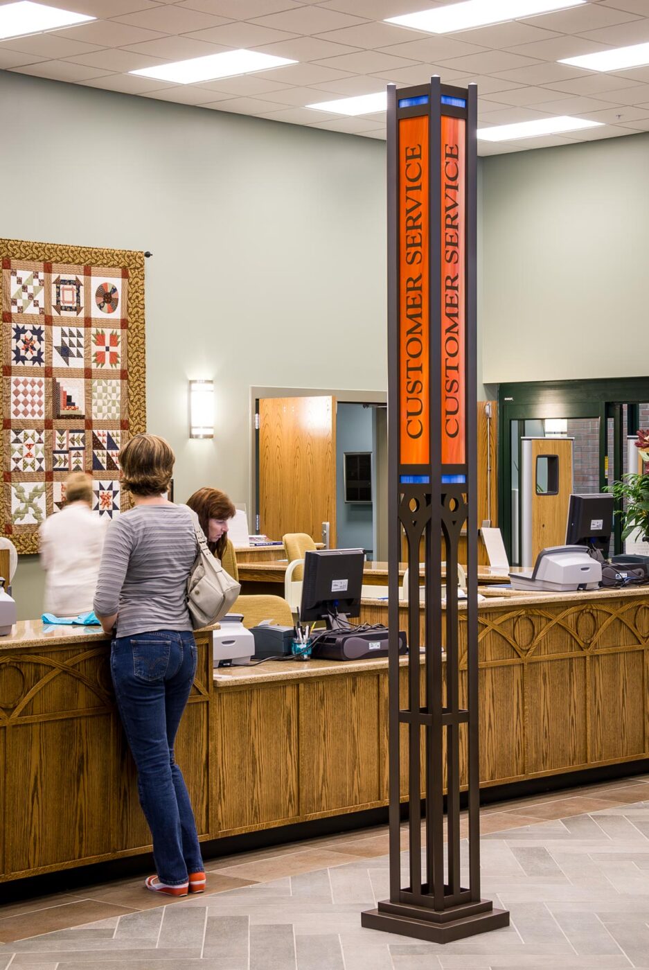 Library interior with tall, thin metal and stained glass customer service sign close the the library counter.
